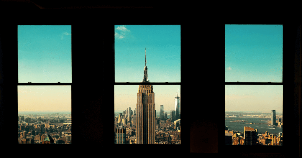 A bright, clean apartment interior with large windows looking out towards a clear view of the Empire State Building in the distance. The room appears tidy and well-maintained.